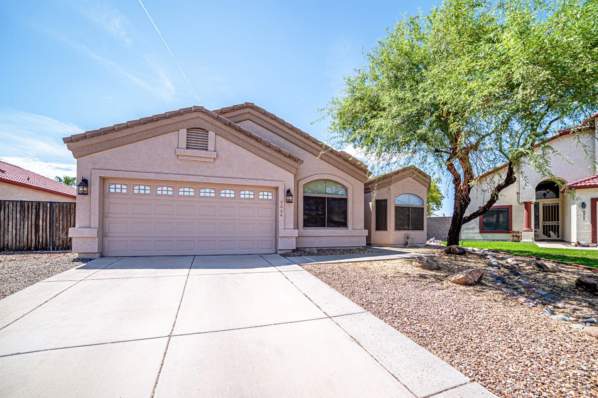 A calm, sunny exterior view of the Phoenix Residential home at Changes Healing Center—featuring beige stucco walls, a tiled roof, two-car garage, desert landscaping, and a shade-giving tree—reflecting a peaceful, homelike setting for recovery and community.