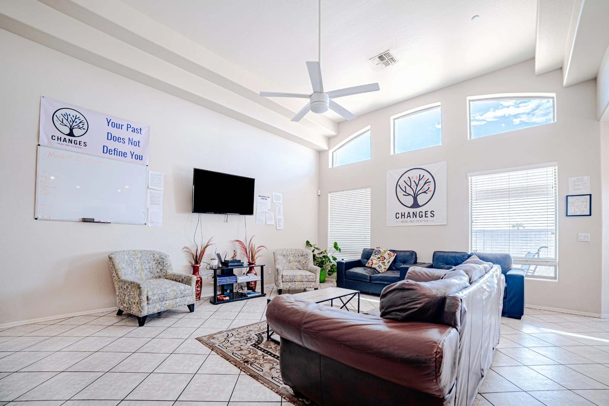 A bright, spacious living room at Changes Healing Center’s Residential Treatment Center in Phoenix—soft natural light floods through high arched windows over a beige-tiled floor. Comfortable sofas and armchairs are arranged around a central coffee table under a ceiling fan, with the Changes logo and the reassuring message ‘Your Past Does Not Define You’ visible on the wall, creating a nurturing and inviting atmosphere.