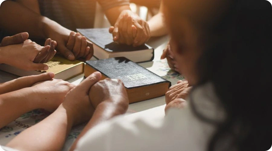 Group of people holding hands in prayer with Bibles on the table at a recovery meeting.