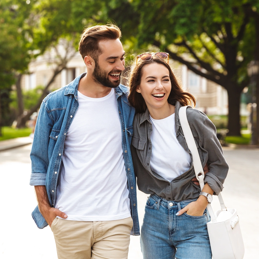 Smiling couple walking arm-in-arm on a sunny, tree-lined street—conveying connection, stability, and renewed well-being.