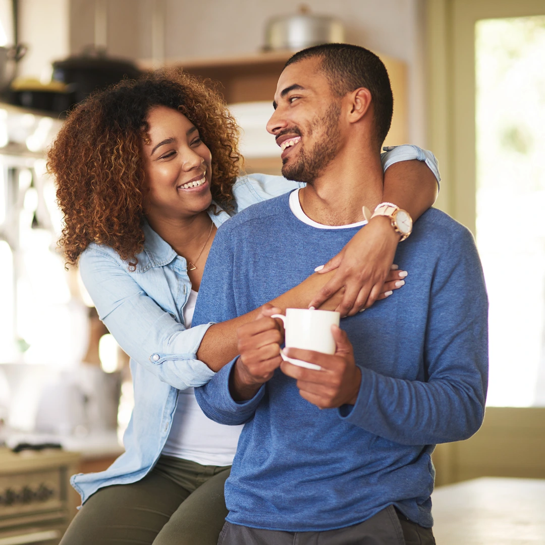Smiling couple embraces in a bright kitchen as one holds a coffee mug, reflecting support during recovery with UnitedHealthcare coverage.