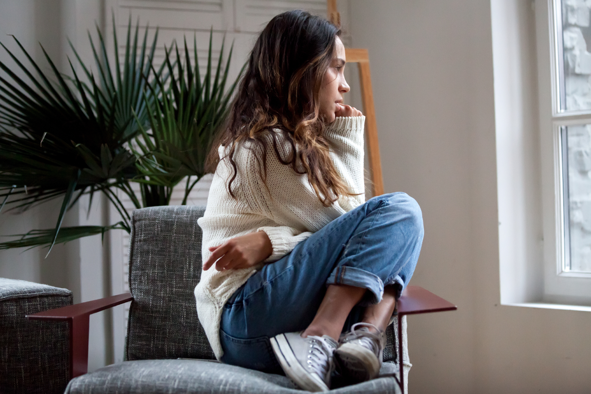 Woman sitting curled up on a chair, looking thoughtfully out a window, reflecting feelings of anxiety or deep contemplation.