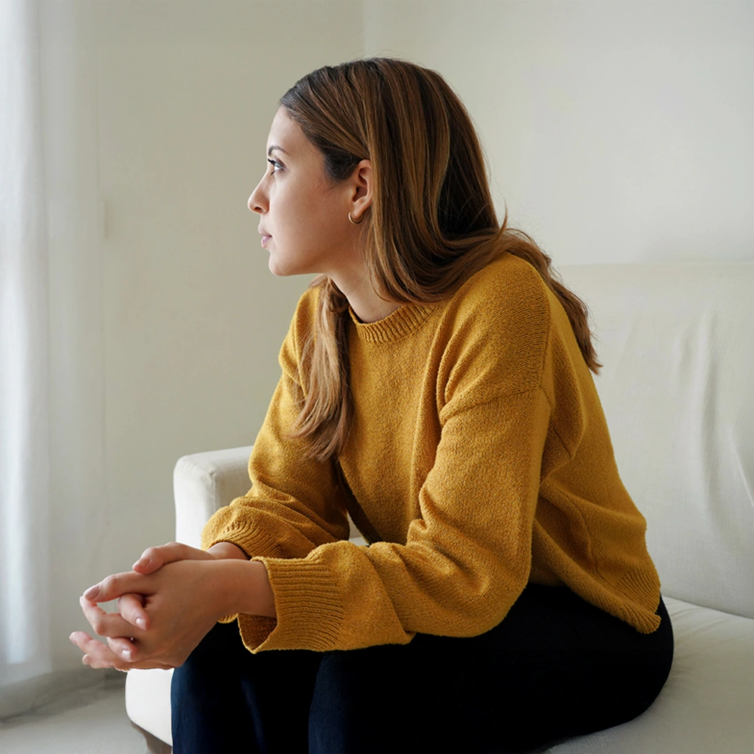 Woman sitting thoughtfully on a couch in a bright room, looking out the window, representing self-reflection and emotional awareness during recovery from codependency.