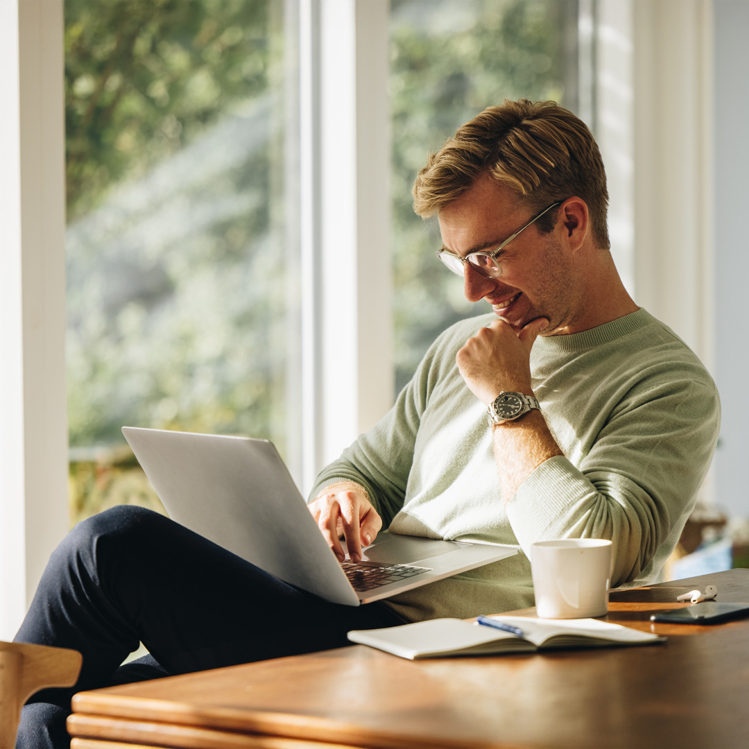 Man sitting by a bright window working on a laptop, smiling thoughtfully with a coffee mug and notebook nearby.