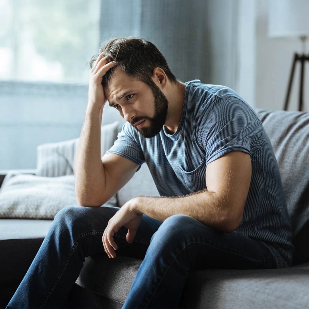 Man sitting on couch with hand on head, appearing deep in thought or distressed, representing emotional challenges of bipolar disorder.