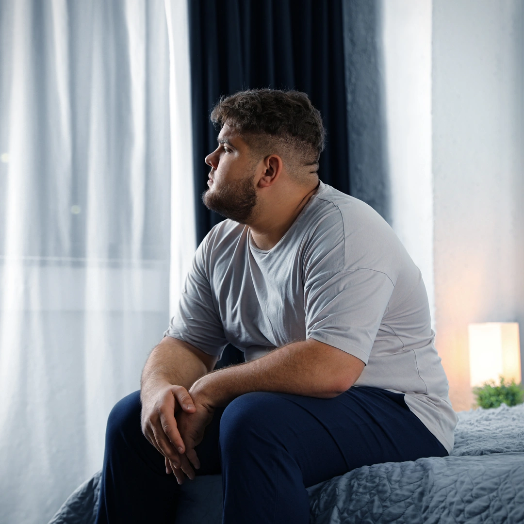 Man sitting on the edge of a bed looking out the window, appearing thoughtful and reflective, symbolizing the emotional challenges and isolation often experienced in codependent relationships.